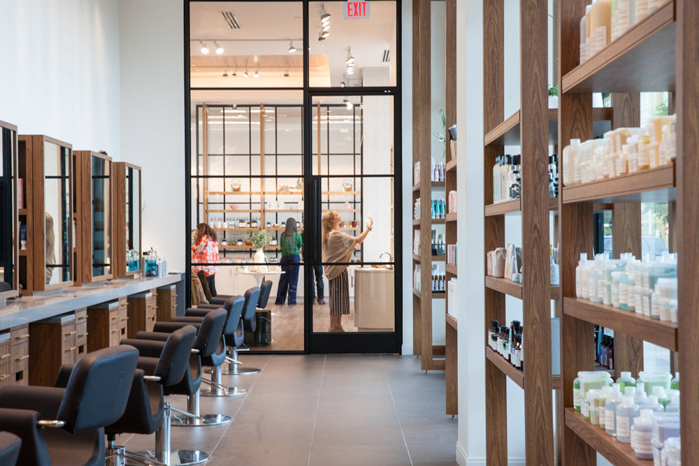 A wide interior view of a modern hair salon with black salon chairs, wood-framed mirrors, shelving filled with bottles, and a glass door looking into another room. Two people are visible near the entrance.