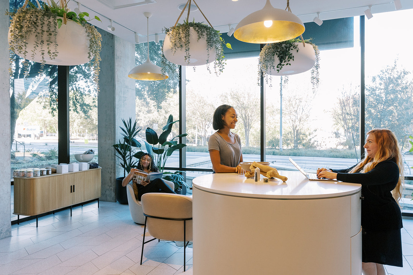A modern interior space with large windows, hanging pendant lights, and several people gathered around a round white reception desk. The scene includes a woman seated on a chair near the window, two standing women at the desk, and decorative plants throughout the room.