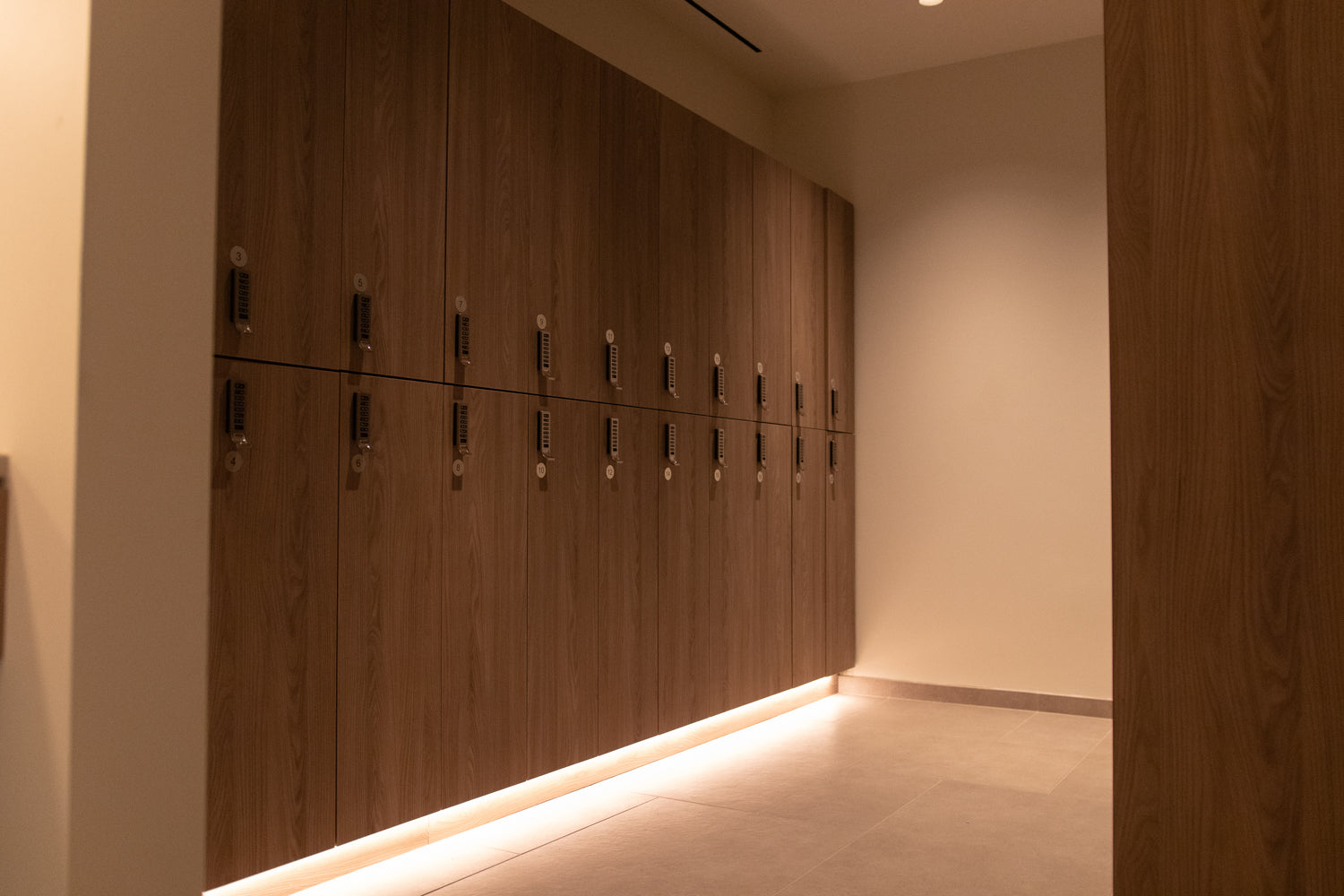 A modern hallway featuring a long wall of dark wood storage cabinets with integrated lockers and keys, illuminated by warm under-cabinet LED lighting.