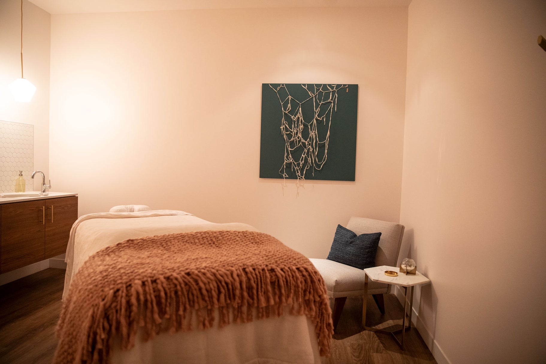 A spa-style treatment room featuring a massage table with a rust-colored fringed throw blanket, a white lounge chair with a blue accent pillow, and a dark teal abstract wall art piece hanging on the beige wall.