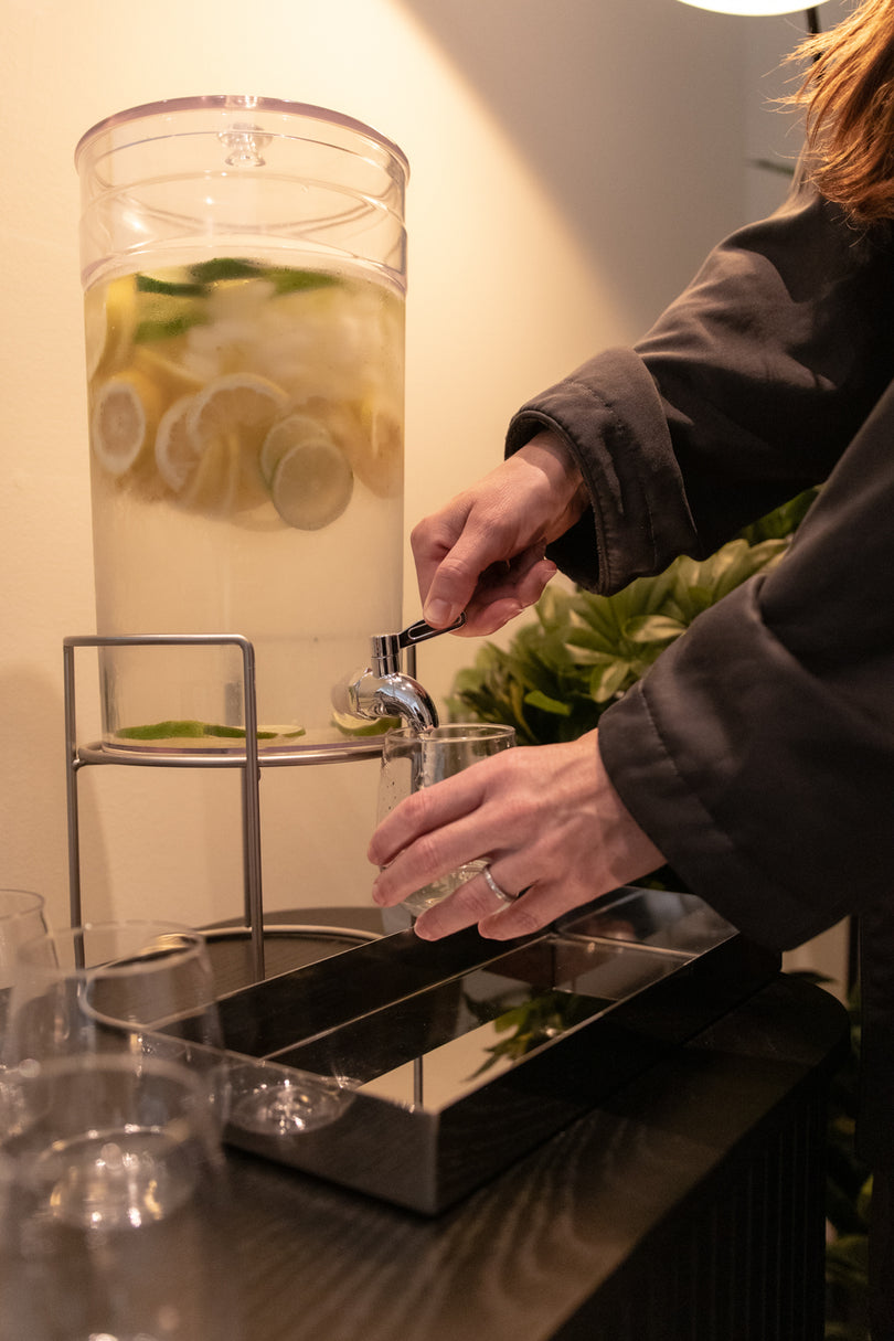 A person in a dark jacket pours a clear glass of water from a large clear beverage dispenser filled with lemon slices and green leaves, set on a dark table.