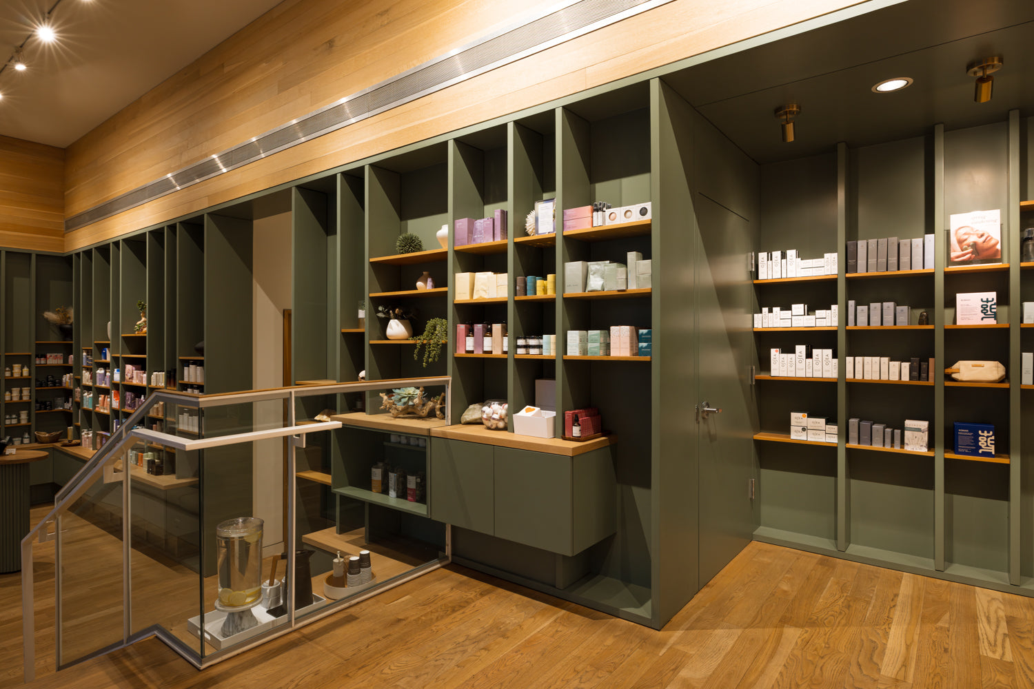 A modern interior retail space with tall built-in shelving in dark green and wood tones, filled with boxed products, set in a warm wood-finished room with a glass railing and ceiling lights.