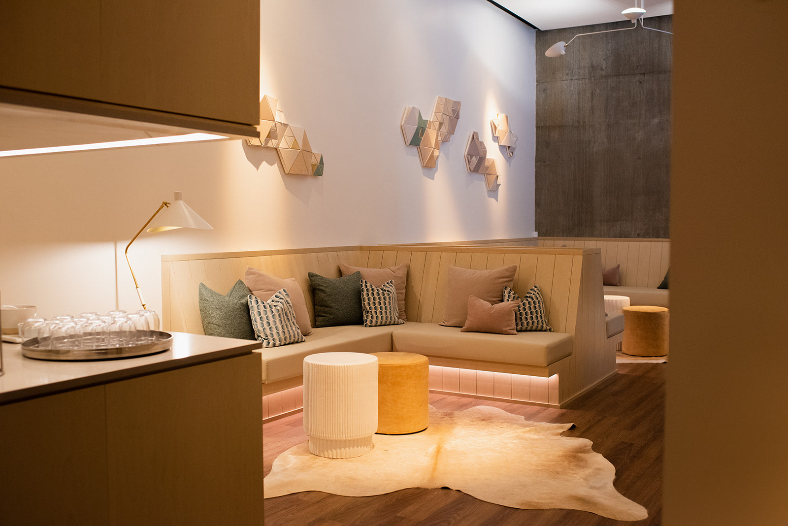 A modern living room corner featuring a beige upholstered corner sofa with multiple accent pillows, a white and yellow cylindrical ottoman, a white cowhide rug, and warm ambient lighting.