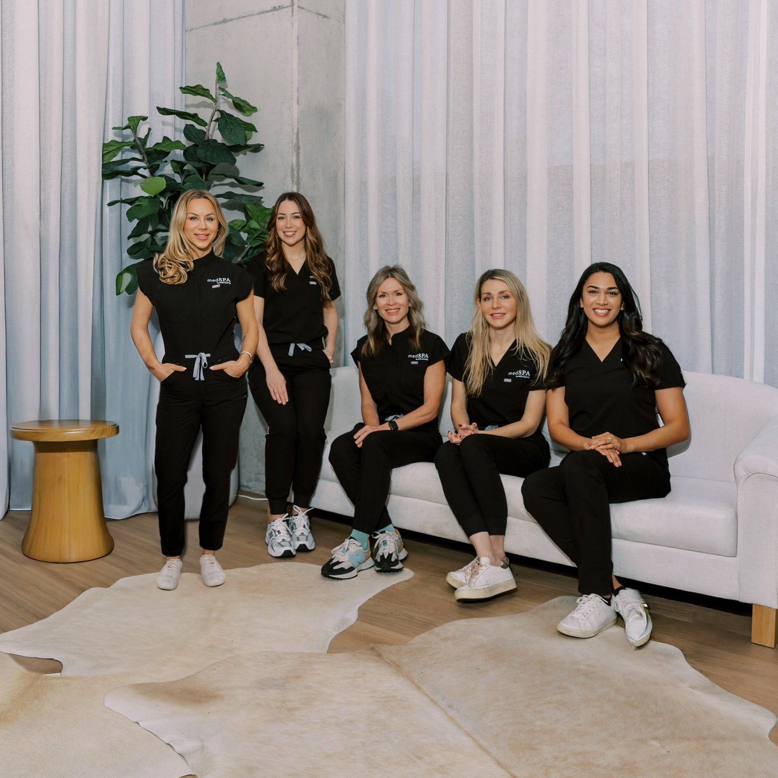Five women in matching black outfits posing in a modern room with white curtains and a white sofa.