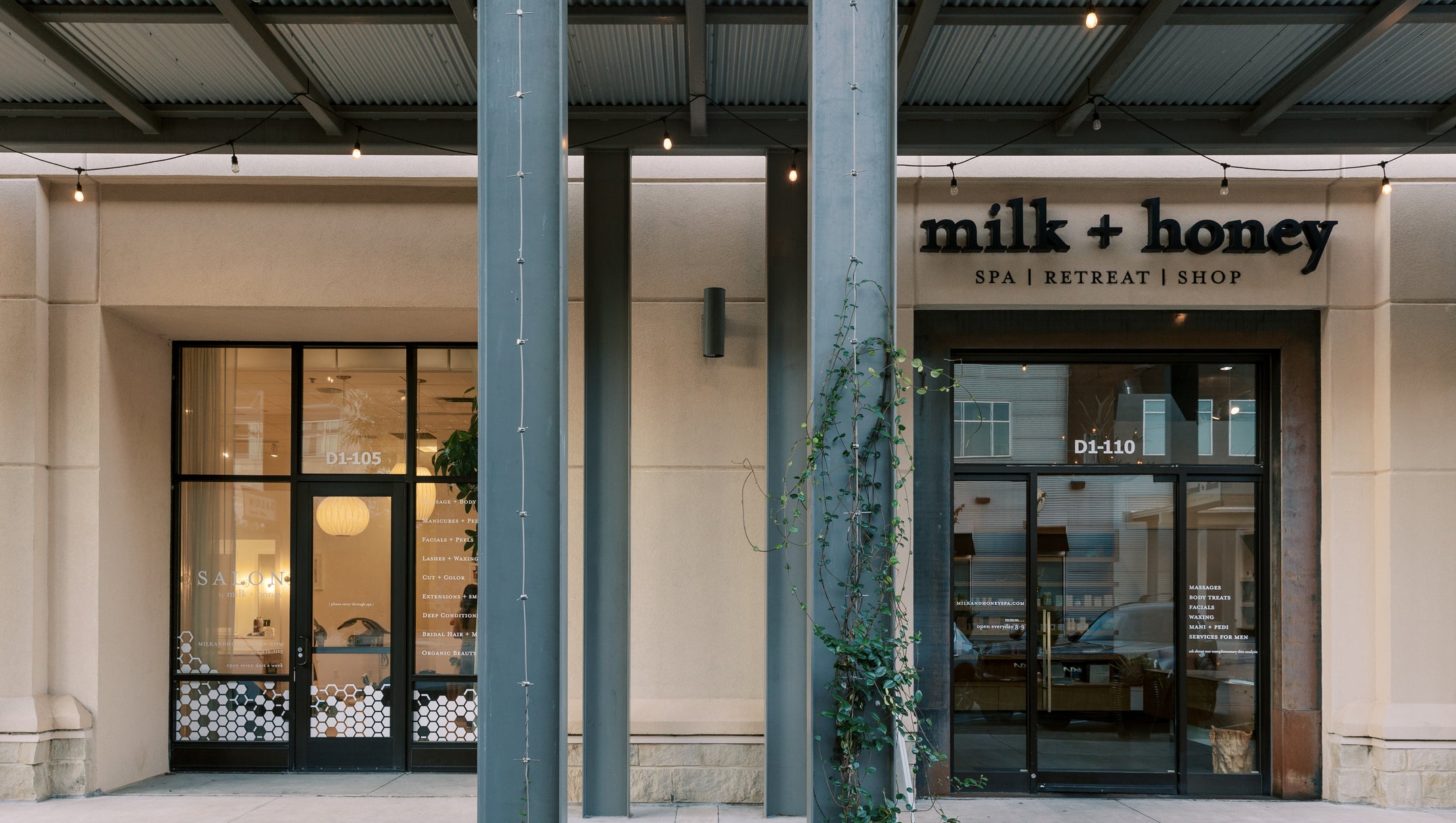 Front exterior of a storefront with the sign 'milk + honey spa | retreat | shop' above glass doors, framed by large gray columns and a covered walkway with hanging lights.