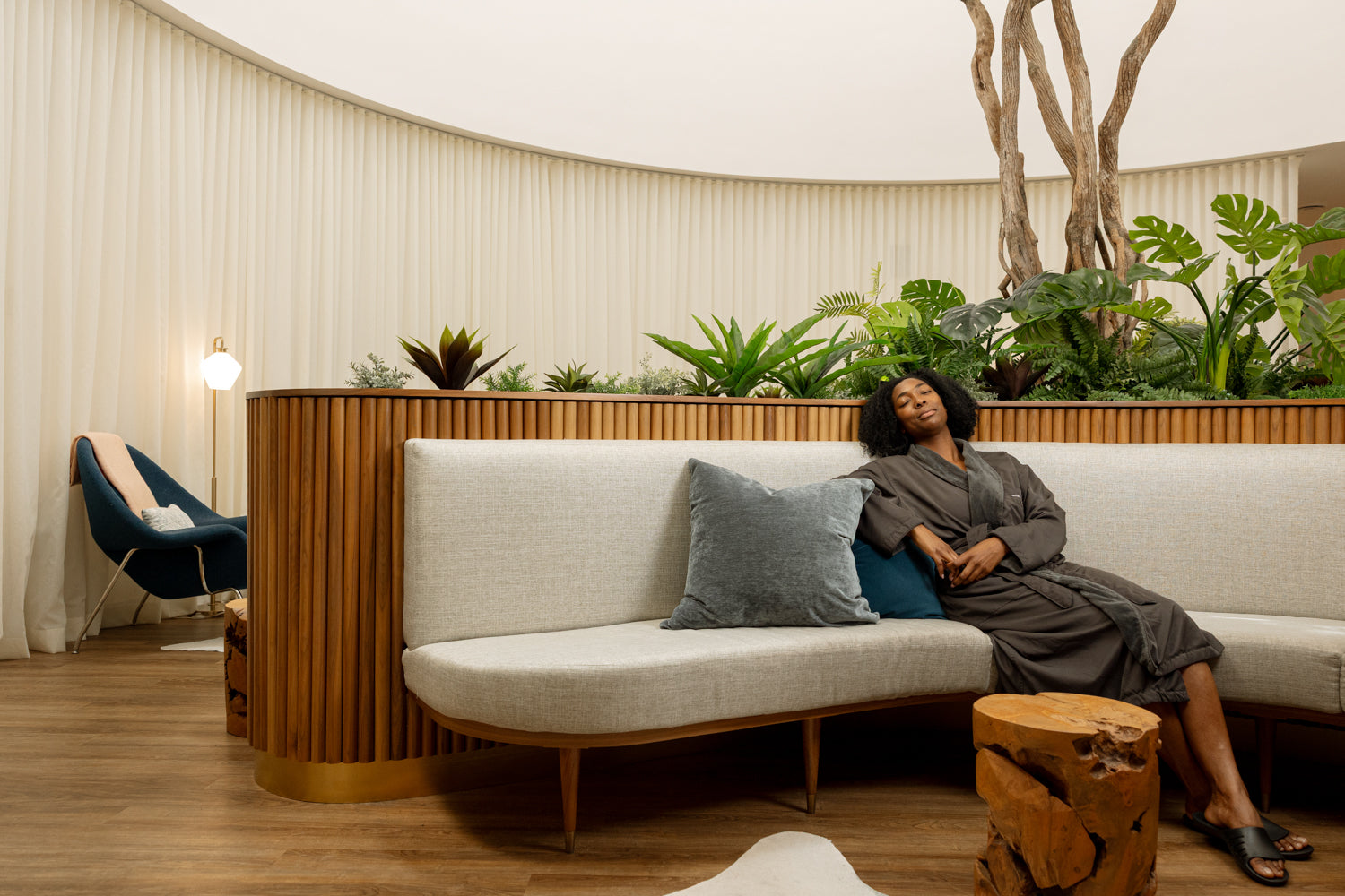A woman in a gray robe reclines on a curved white sofa with a blue pillow in a modern indoor lounge setting with wood slat paneling, plants, and a tree trunk side table.