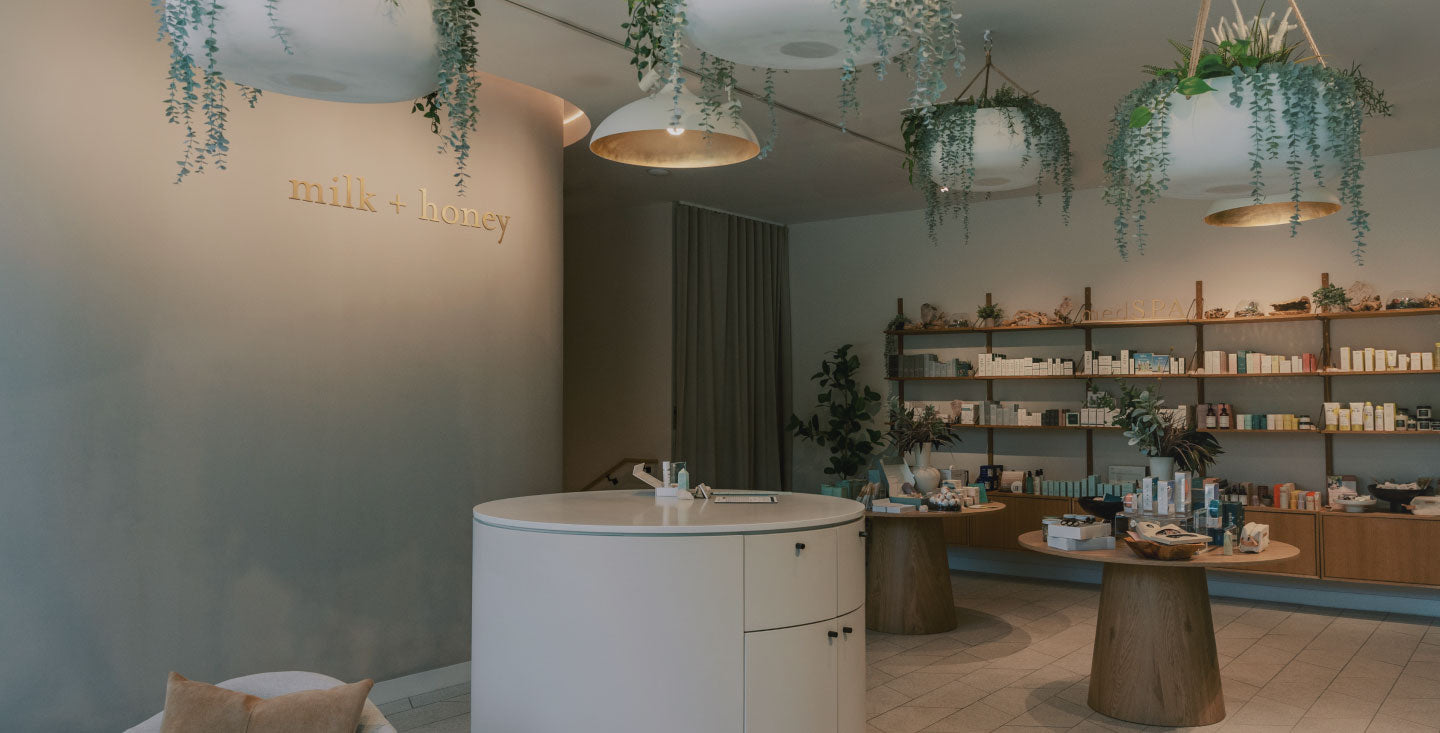 A modern skincare or beauty retail interior with a round white counter, wooden display tables, shelves of products, hanging greenery, and warm pendant lights. The wall displays the brand name 'milk + honey' in gold lettering.