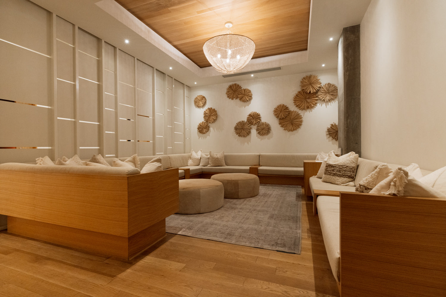 A modern, warm-toned lounge or spa interior with wooden seating, cream cushions, a round glass chandelier, wood ceiling panels, and decorative woven wall pieces.