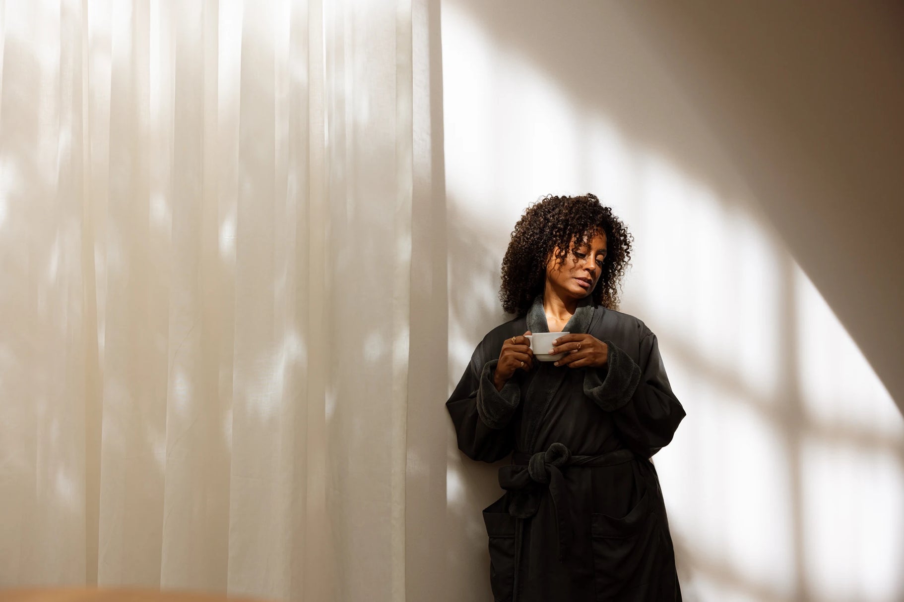 Person in a black robe holding a cup against a wall with light filtering through.