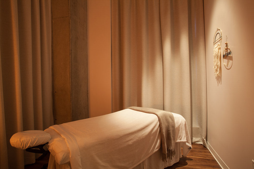 A massage table covered in white linens with a beige throw blanket, set in a warmly lit spa room with draped curtains and wood flooring.