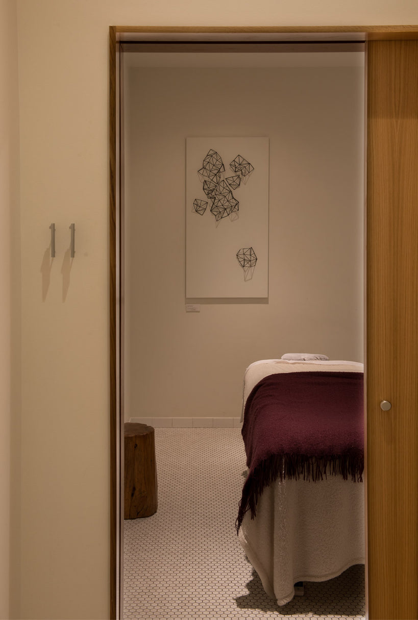 A view through a doorway into a spa or treatment room, showing a massage table with a deep purple throw blanket, a small wooden stool, and a framed abstract black line drawing on the wall.