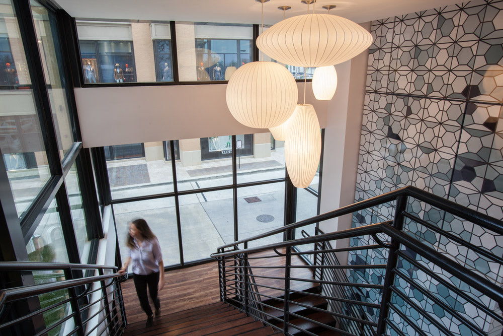 A modern interior staircase with black metal railings, wooden steps, large white ribbed pendant lights, and a patterned wall on the right. A woman is walking down the stairs near the left side.