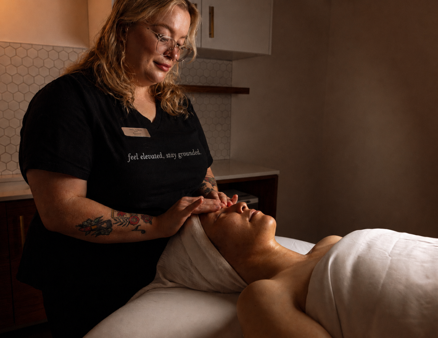 A spa therapist in a black scrub top with white text applies a facial treatment to a client lying on a treatment bed in a warmly lit spa room.
