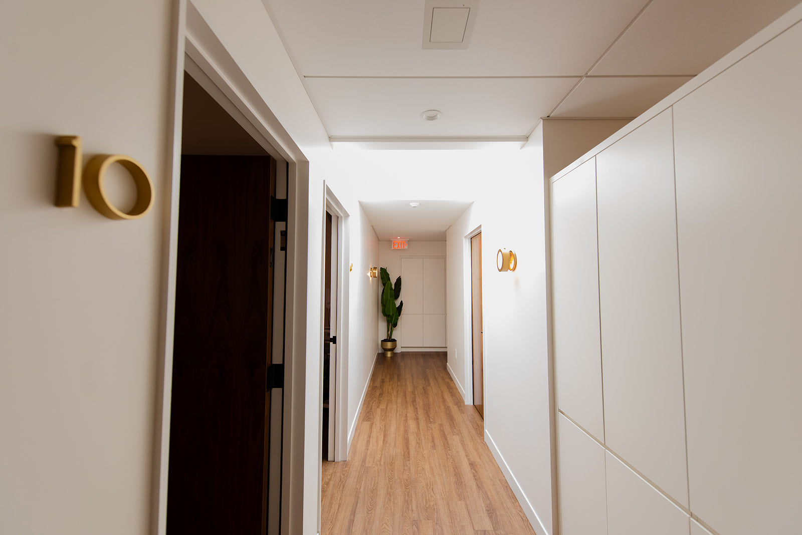 A modern hallway with light wood floors, white walls, recessed ceiling lights, a small potted plant in the distance, and gold room-number wall decor.