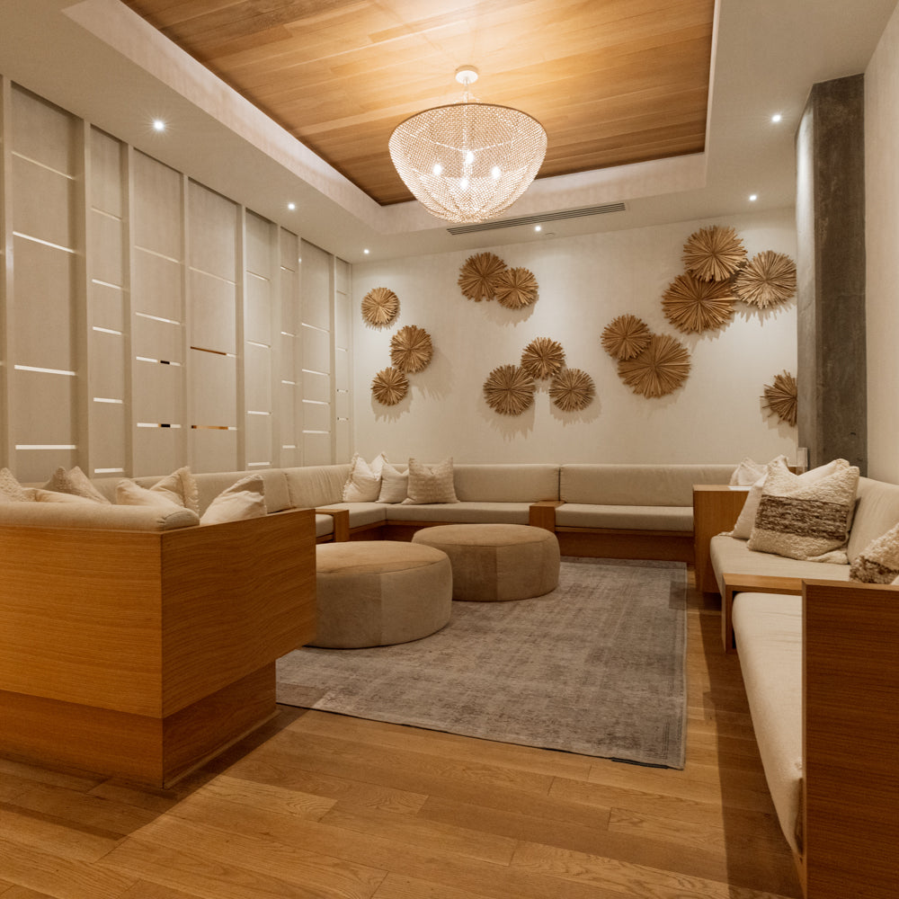 A modern, warm-toned lounge or spa interior with wooden seating, cream cushions, a round glass chandelier, wood ceiling panels, and decorative woven wall pieces.