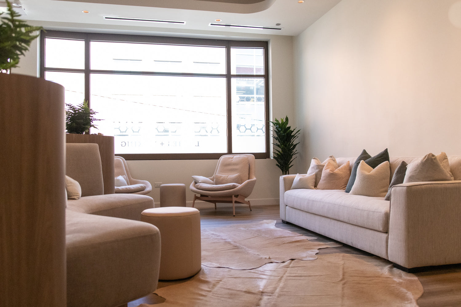 A modern living room featuring a light beige sectional sofa with matching throw pillows, a matching beige armchair, a round ottoman, and a large cowhide rug on a wood floor, with a large window in the background.