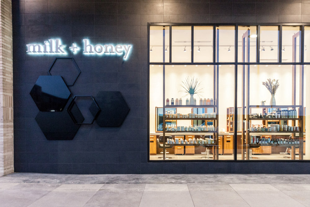 Milk + Honey storefront exterior with a dark gray wall, glowing white neon-style logo, black hexagonal wall decor, and large glass windows showing shelves inside.