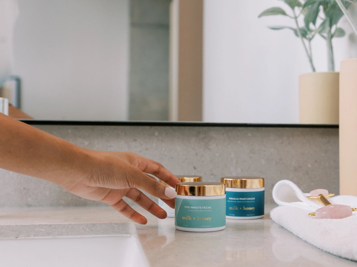 Hand reaching for a skincare product on a bathroom counter with a mirror and towel in the background.