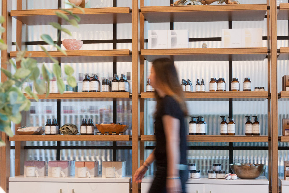 A woman walks past tall wooden shelves stocked with small bottles, jars, and boxes in a bright retail interior.