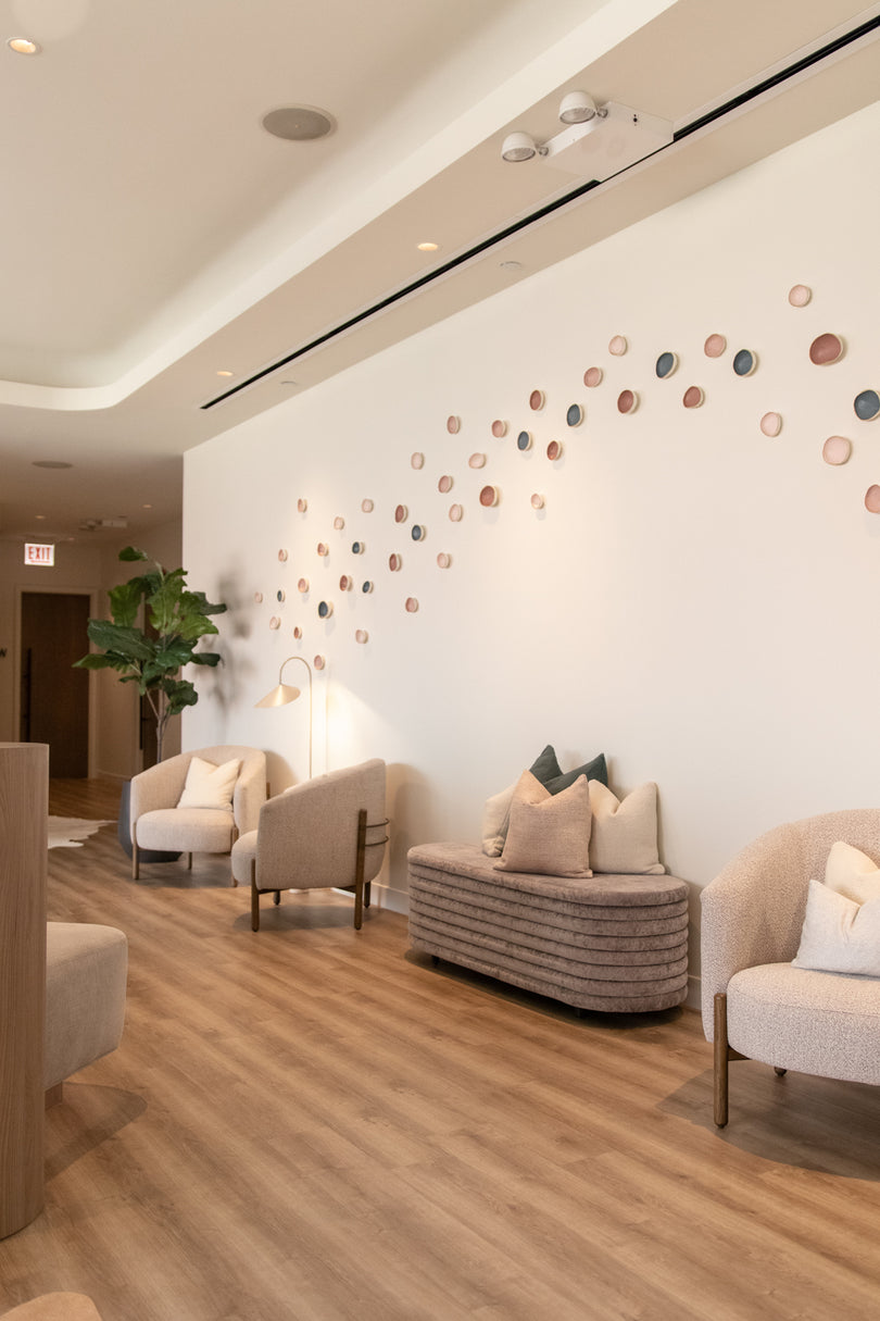 A modern interior lounge or waiting area with light wood flooring, cream upholstered armchairs, a ribbed bench with pillows, and a white wall decorated with many small circular wall pieces in muted pink, beige, and gray tones.