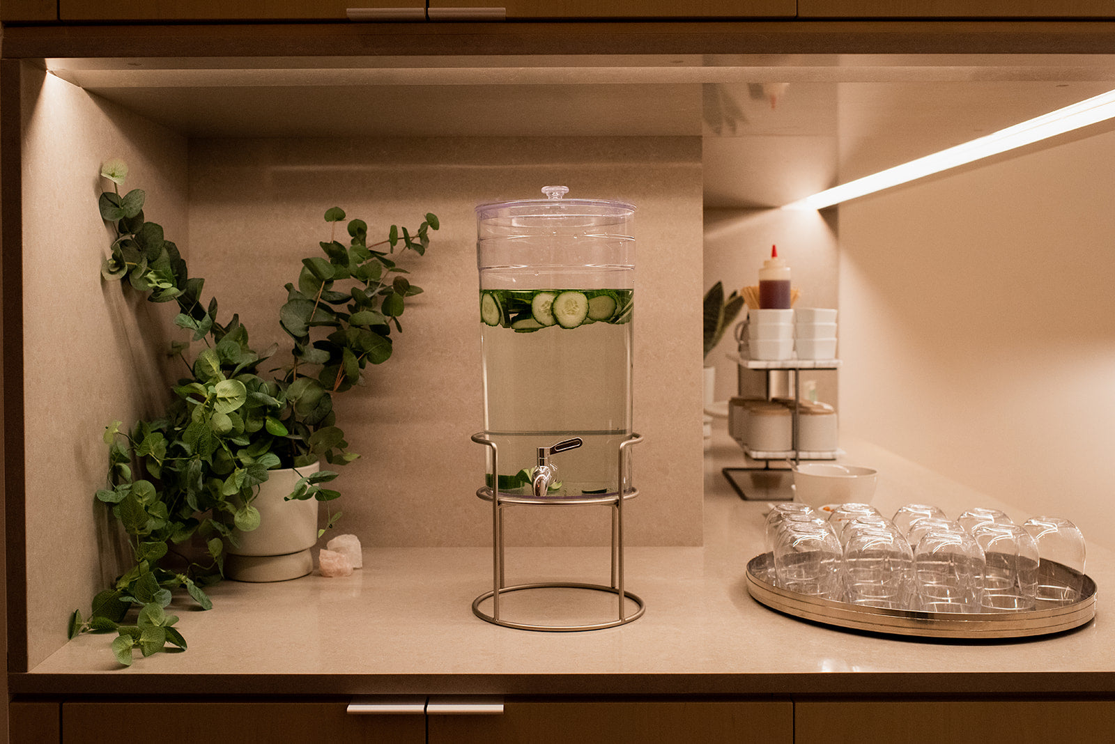 A clear glass water dispenser with a metal stand, filled with water and cucumber slices, set on a beige countertop in a warm, modern kitchen or bar area with plants and glassware.