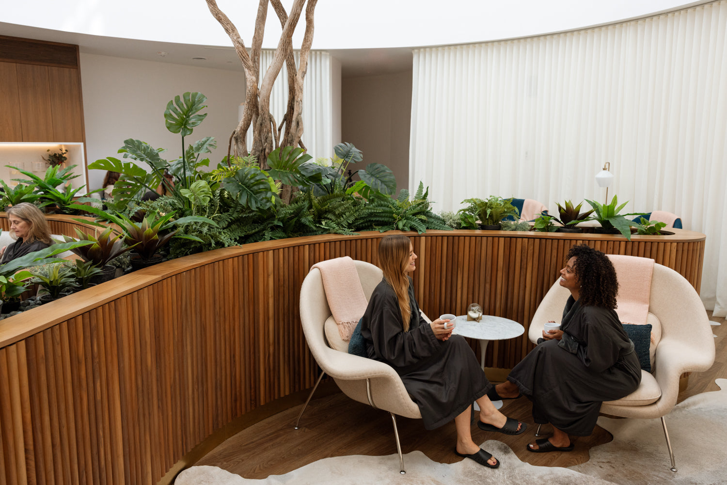 Two women in black robes sit in cream-colored lounge chairs with a small round table between them, inside a modern indoor garden with curved wooden slat walls, lush green plants, and white curtains in the background.