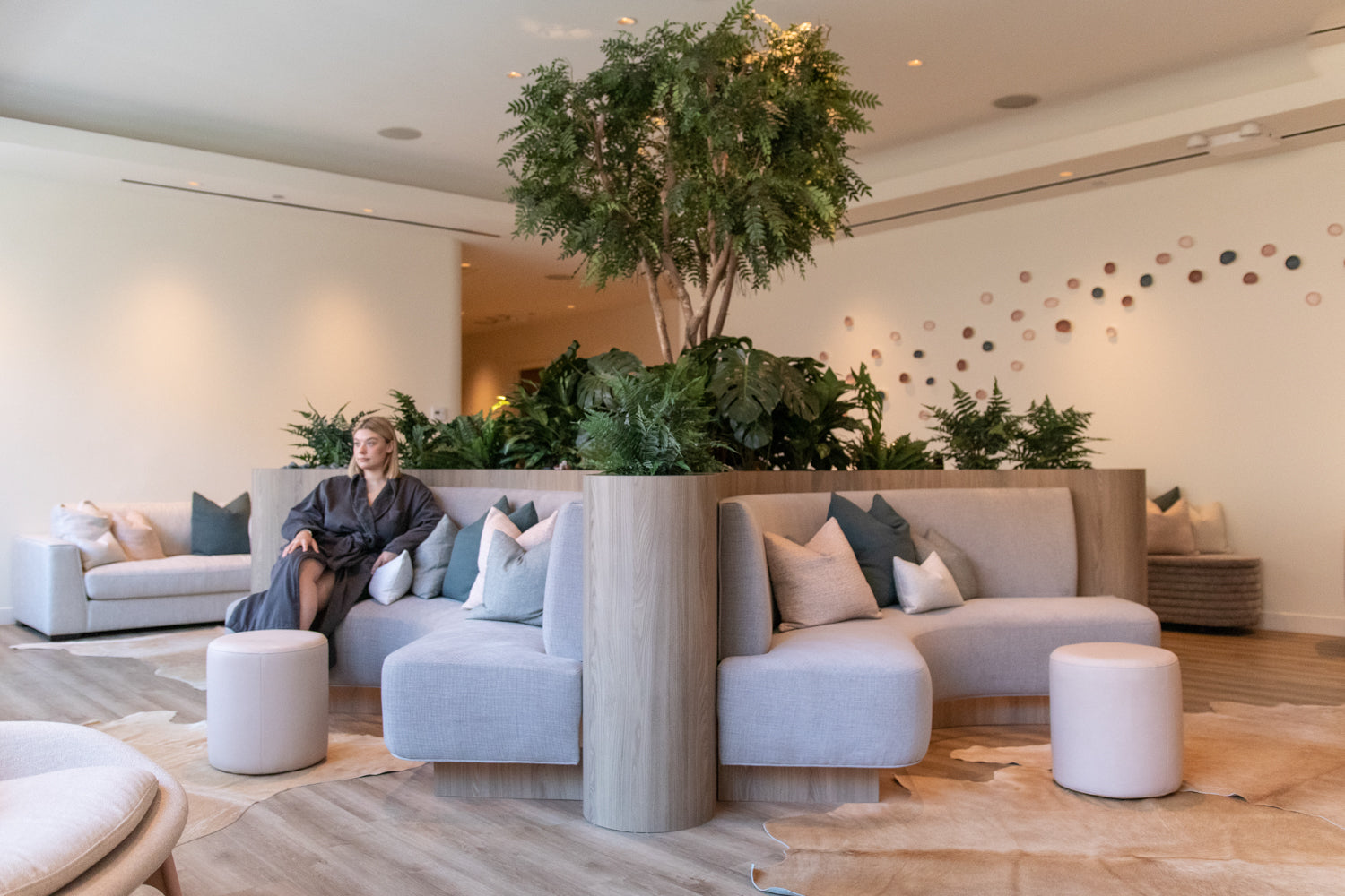 A woman in a dark robe sits on a light gray modular lounge sofa in a bright, modern spa-like interior with wood floors, potted greenery, and matching poufs.