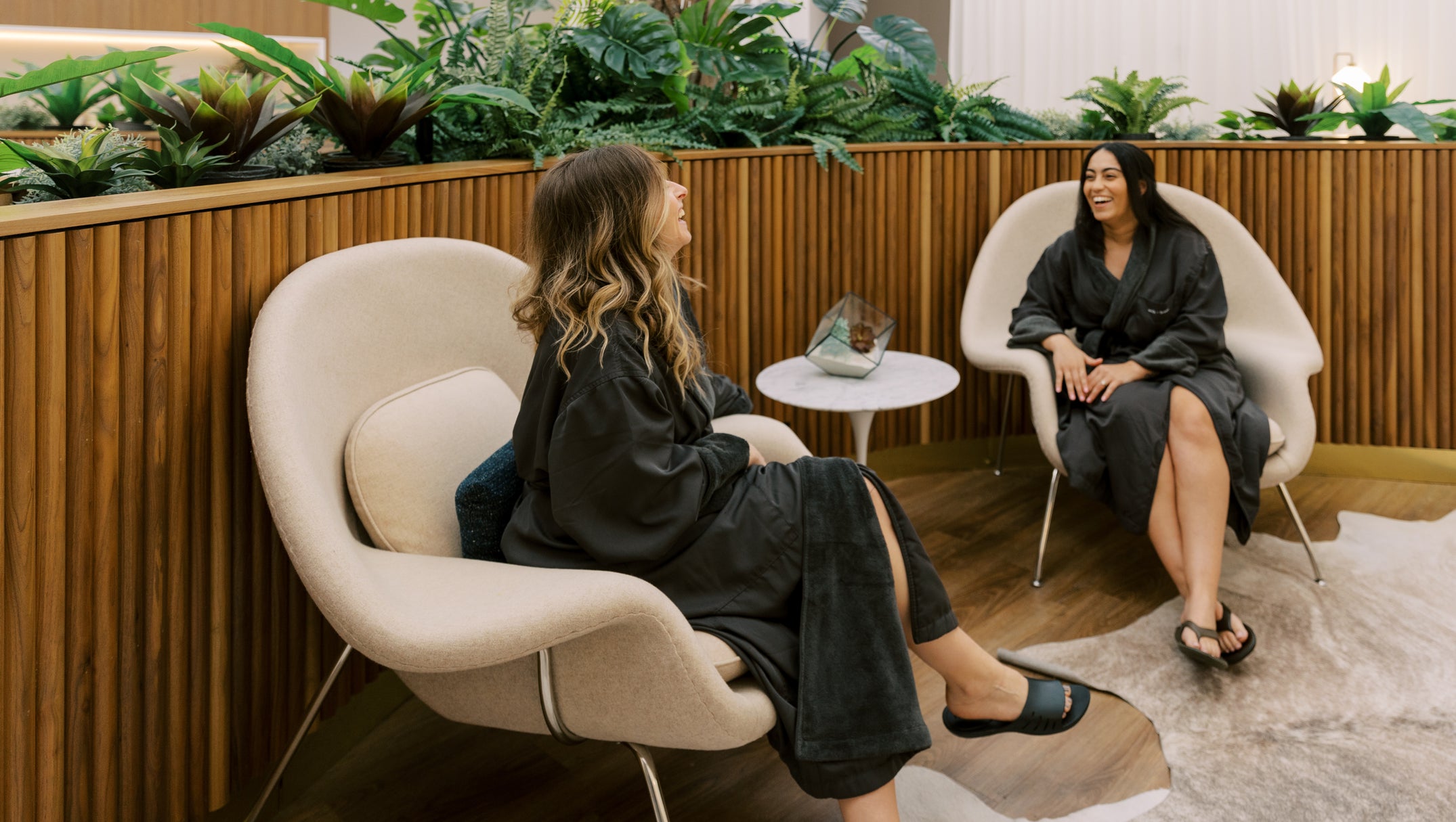 Two women in black robes sitting in modern chairs with plants in the background