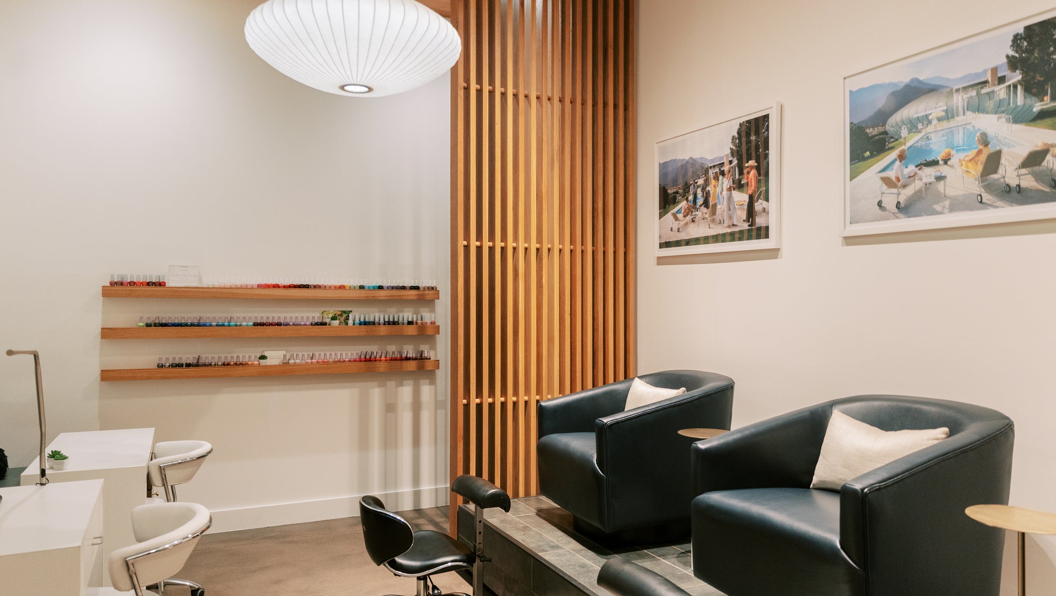 A modern spa or salon interior with two black leather reclining chairs, a pedicure treatment table, white vanity tables, wall shelves, framed artwork, and a slatted wood screen.