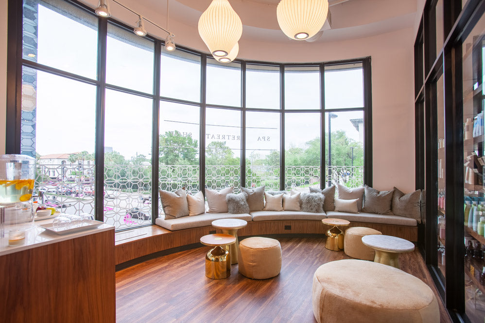 A modern spa-style interior with a curved upholstered bench, round ottomans, small round tables, large black-framed windows, and pendant lights. No person or animal is present, and the scene appears to be a real environment rather than a clean isolated product shot.