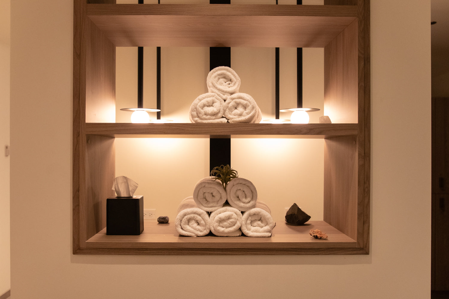 A built-in wooden wall shelf with warm lighting, styled with neatly rolled white towels, a tissue box, and small decorative objects against a beige wall.