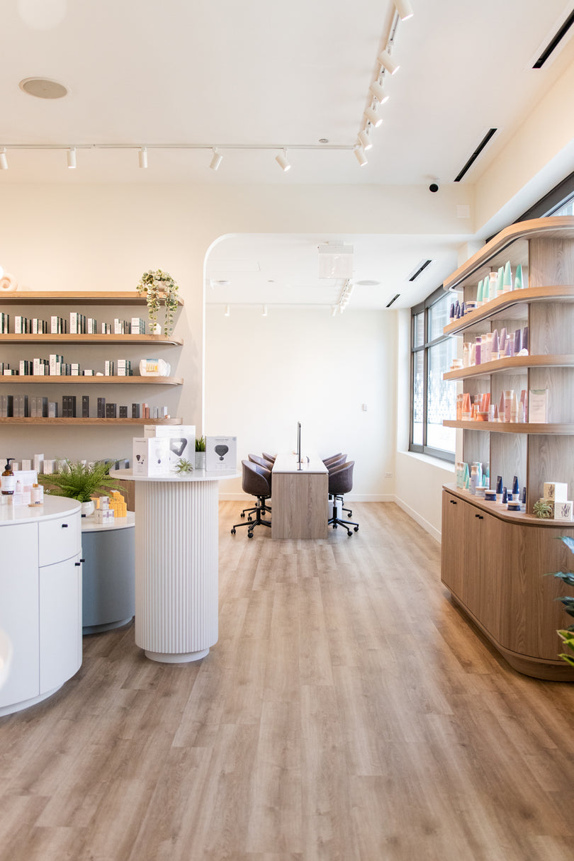 A bright, modern spa or beauty salon interior with light wood flooring, white and wood shelving filled with products, a central white pedestal counter, and two treatment chairs in the distance.
