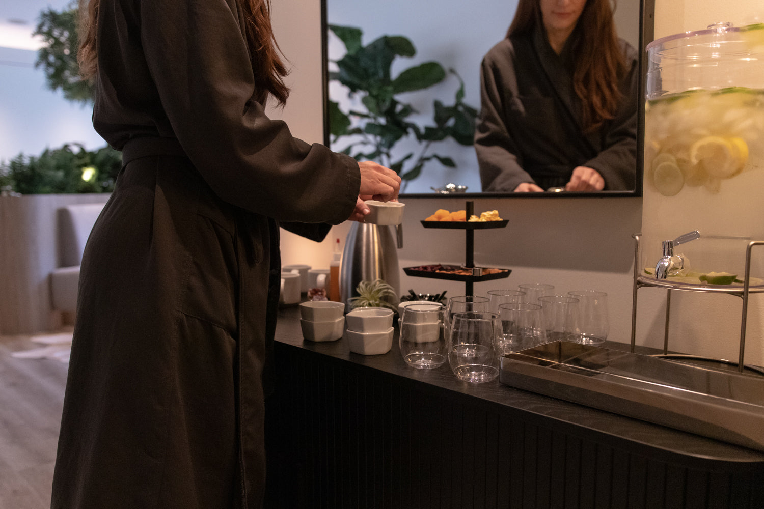 A woman in a dark brown robe stands at a beverage station, pouring from a metal kettle into glass cups. A large mirror reflects her, and a lemonade dispenser is visible on the right.