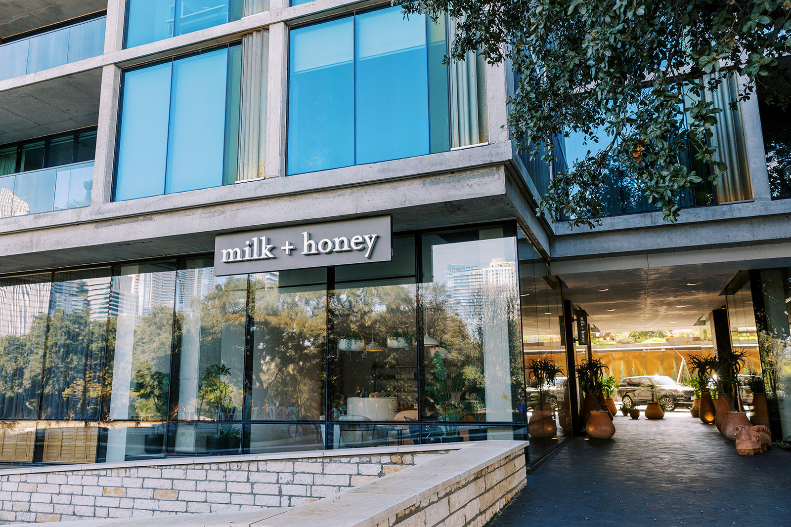 Modern glass-front building exterior with a visible 'milk + honey' sign above the entrance, reflections of trees in the windows, and a paved walkway leading into the lobby.