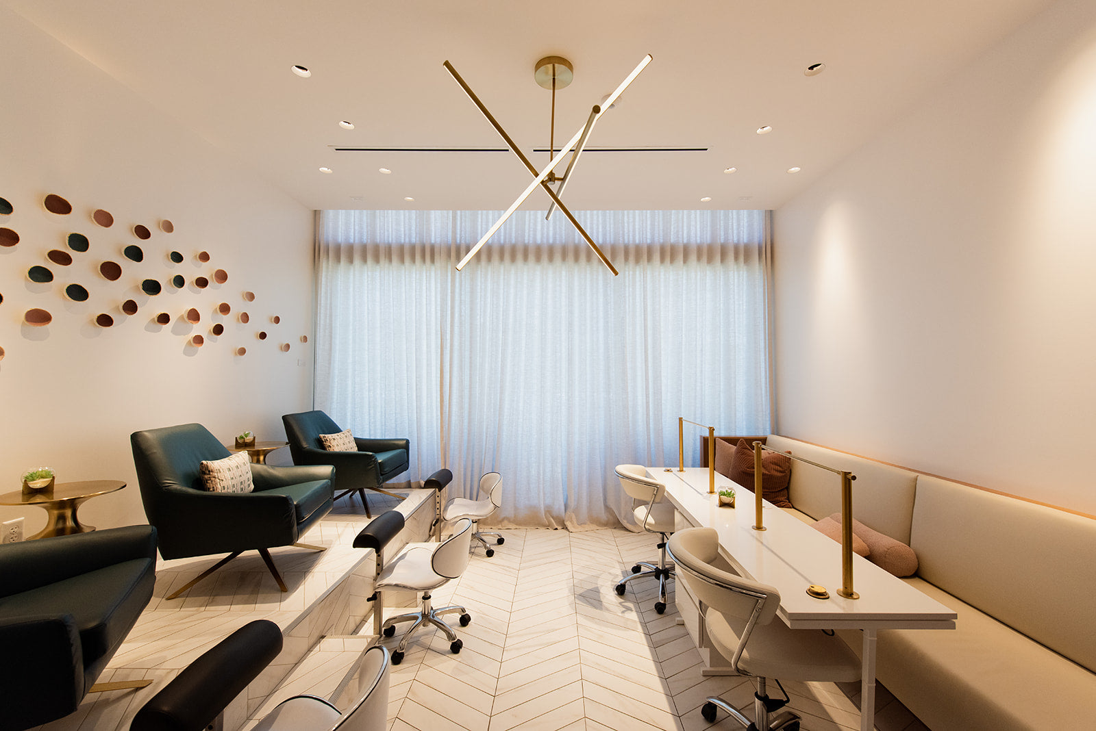 A modern, bright spa or salon interior with teal lounge chairs, white rolling office chairs, a long cream-colored reception desk with gold accents, and a large window covered in sheer white curtains.