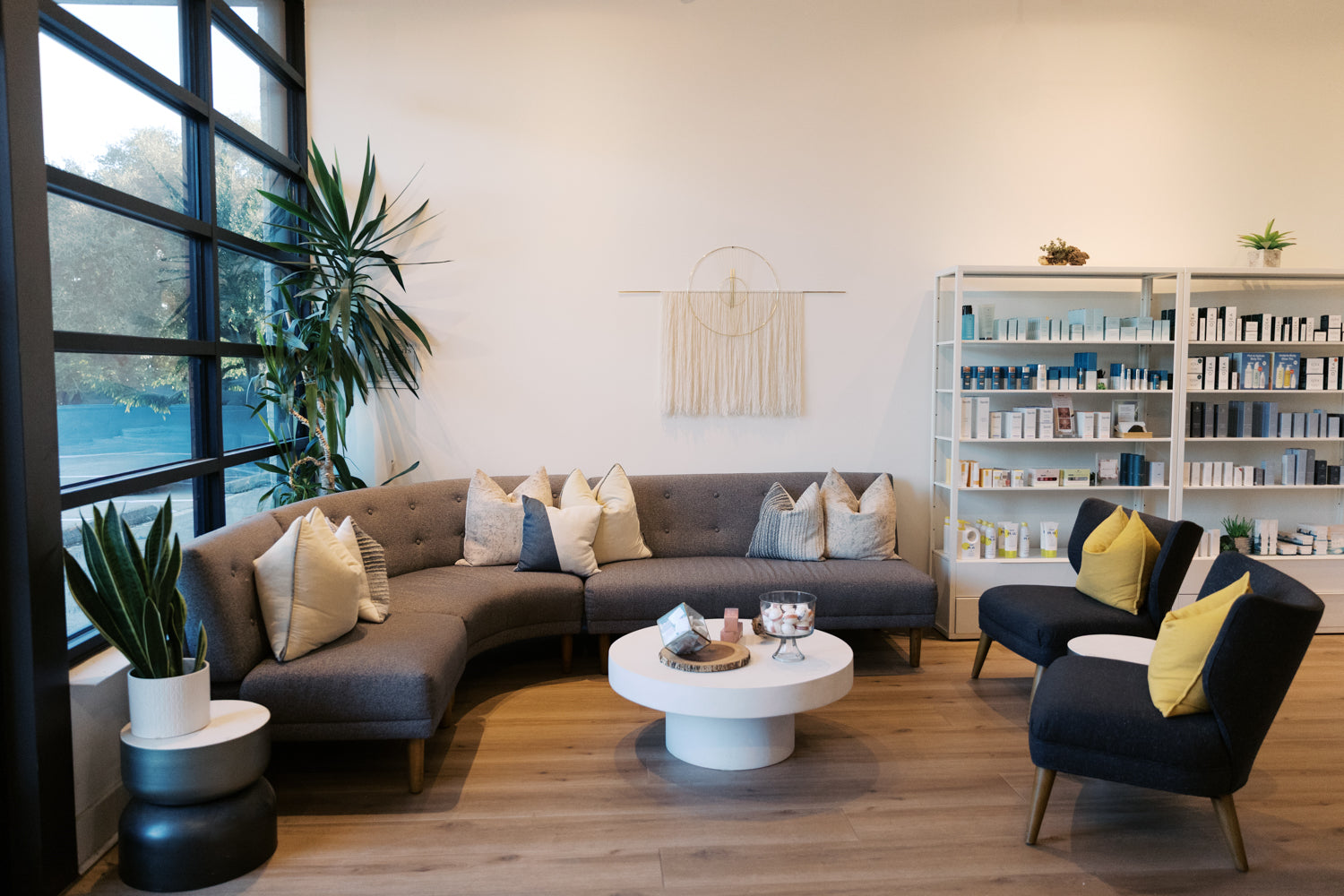 A modern living room setup featuring a curved gray fabric sofa with beige and blue-gray pillows, a round white coffee table, two dark blue armchairs with yellow pillows, a white shelving unit filled with products, and a large window with black frames.