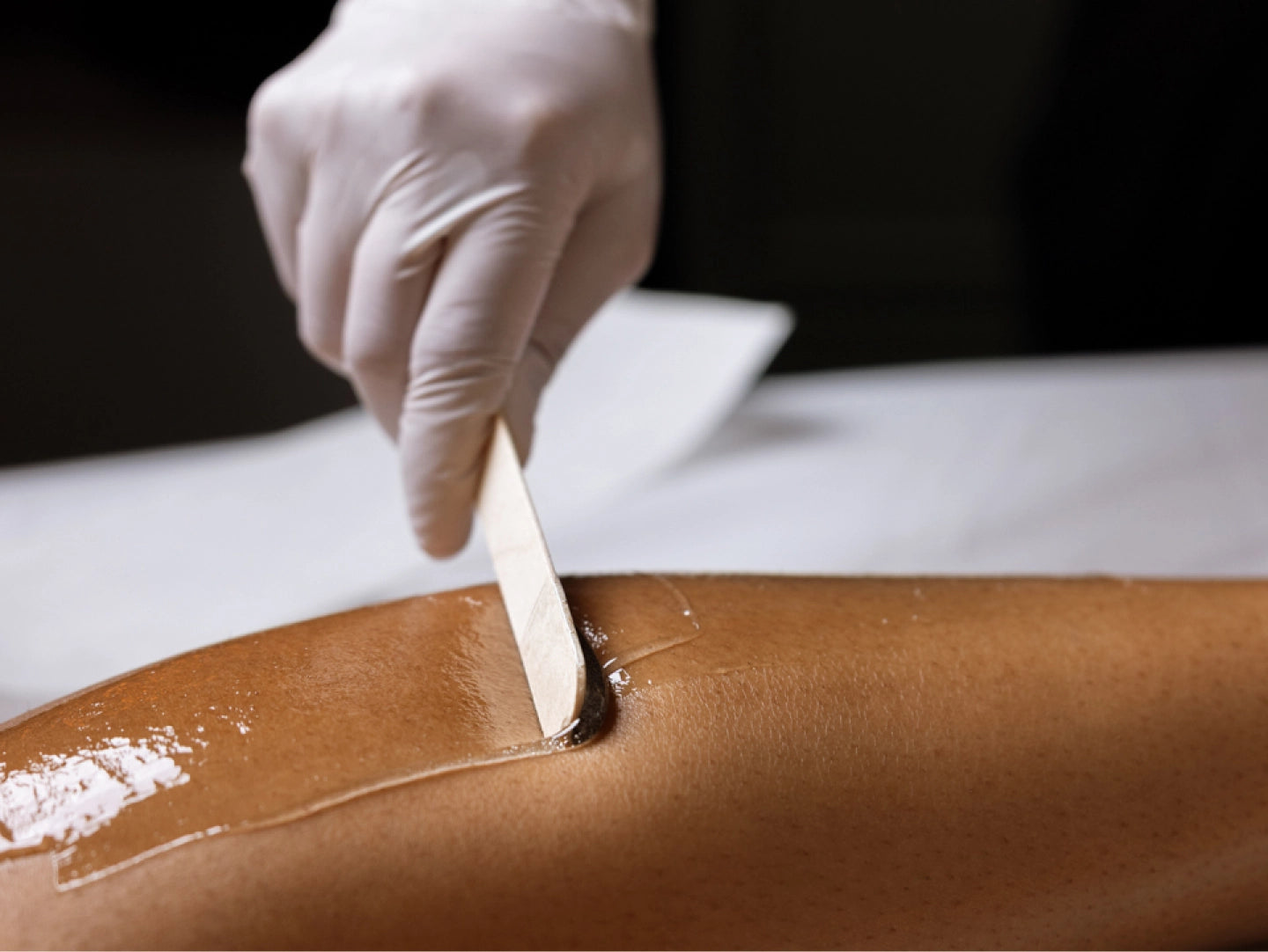 Person applying brown paste to a surface with a wooden spatula, wearing white gloves.