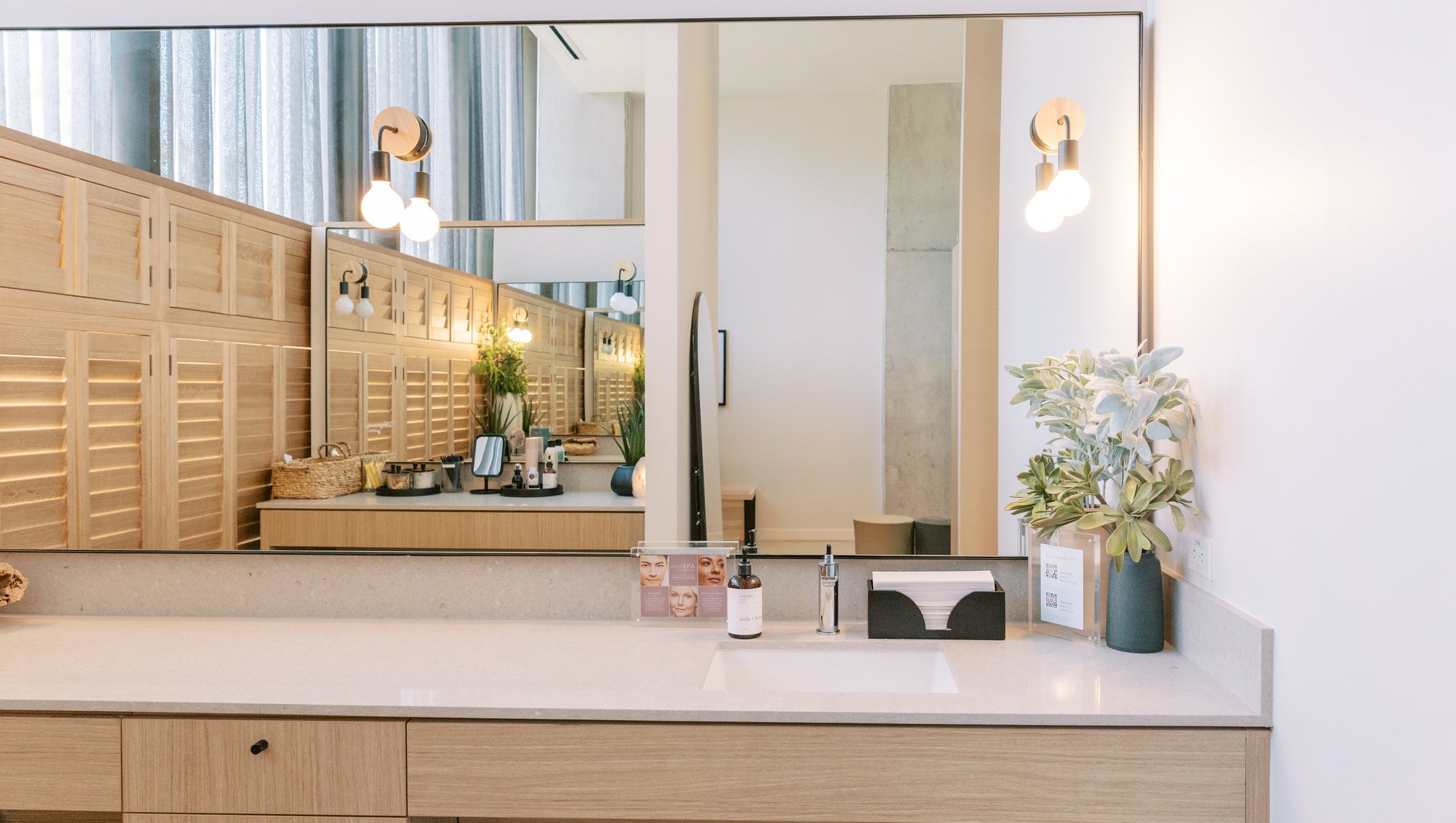 A modern bathroom vanity with a light wood console table, a white countertop, a large wall mirror, and warm wall sconces. A vase with white flowers sits on the right side of the counter.