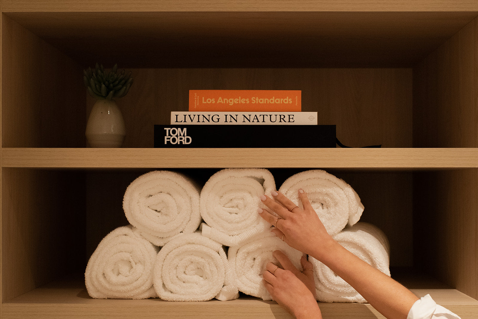 A pair of hands arranging white rolled towels on a wooden shelf, with books and a small vase displayed on the upper shelf.