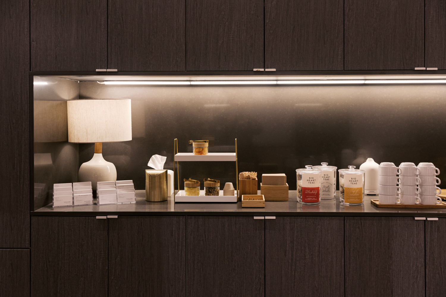 A modern beverage and snack bar counter set against dark wood cabinetry, featuring a white ceramic table lamp, stacks of paper cups, small jars, and boxes under warm under-cabinet lighting.
