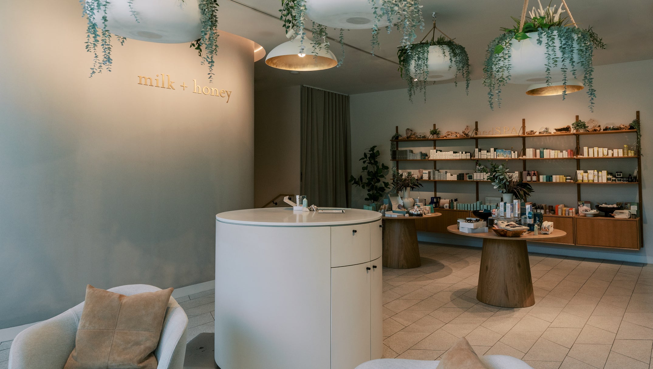 A modern spa or beauty retail interior with a round white reception counter, hanging planters, shelving filled with products, and neutral-toned seating. The wall displays the text 'milk + honey'.