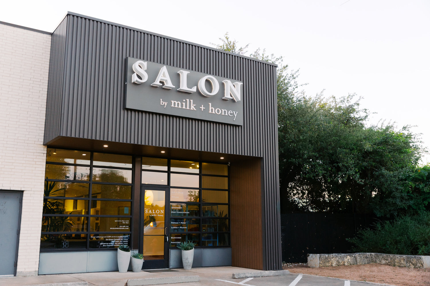 A modern storefront with a dark gray corrugated metal facade and a large sign reading 'SALON by milk + honey' above glass entrance doors, with trees and a pale sky in the background.