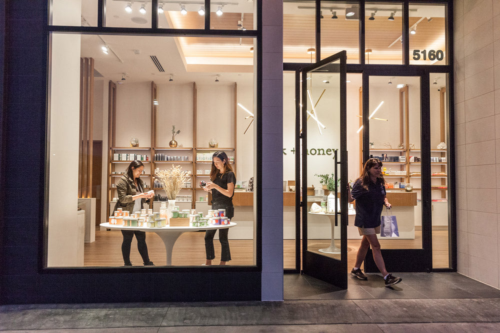 A storefront window display with large black-framed glass doors and windows, showing a warmly lit interior shop. Two women stand inside near a small display table, and one woman walks out through the open door.