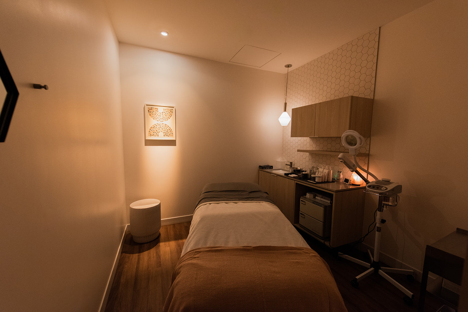 A compact spa or massage room with a single treatment bed, warm ambient lighting, wood flooring, wall cabinets, and a small vanity area with a fan and mirror.
