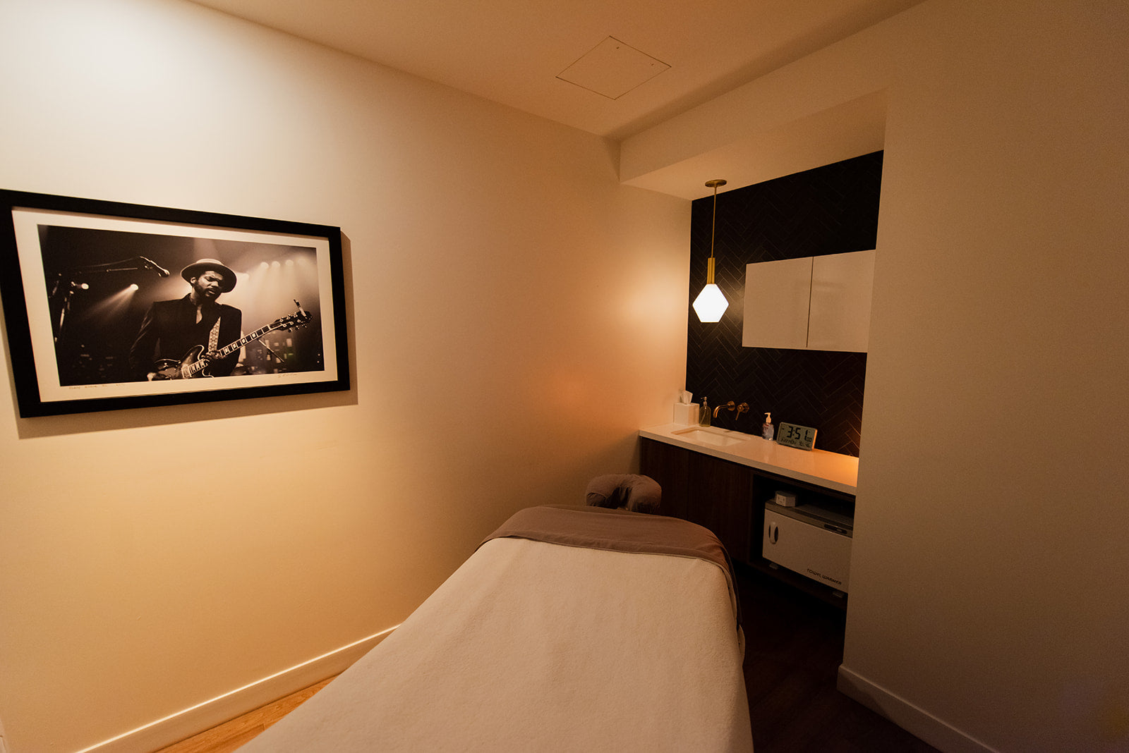 A spa treatment room featuring a massage table in the foreground, a black-framed black-and-white photograph on the beige wall, and a modern vanity area with a hanging pendant light in the background.