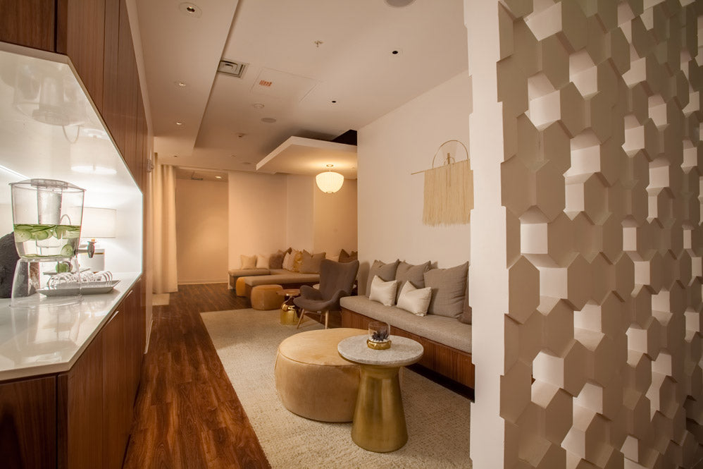 A modern interior space with a beige upholstered sofa, round ottoman, small round coffee table, wood flooring, and a decorative geometric wall panel on the right.