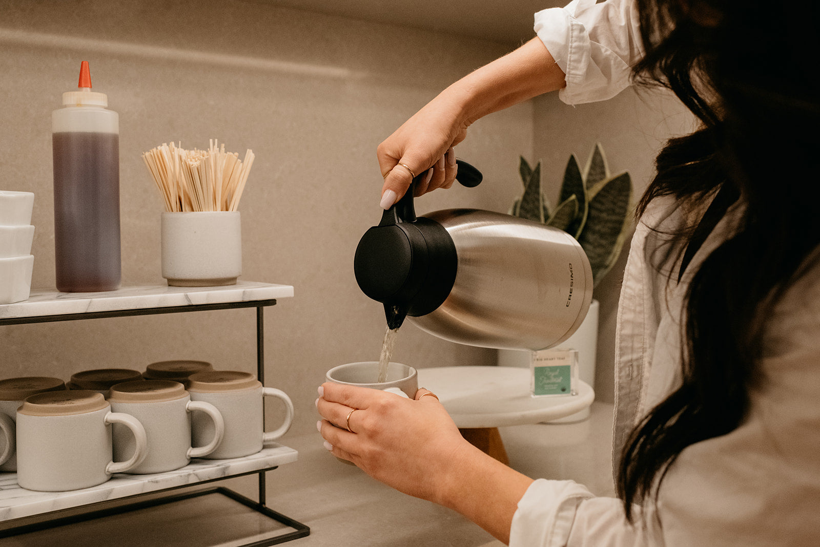 A person pouring hot water from a stainless steel kettle with a black lid into a white mug on a shelf in a kitchen or cafe setting.