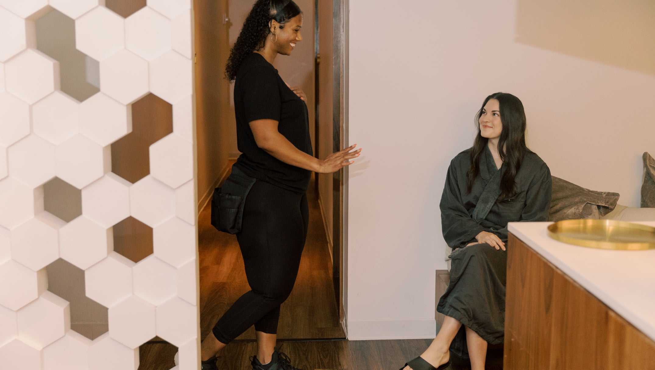 Woman in black outfit entering a room where another woman is sitting on a couch.