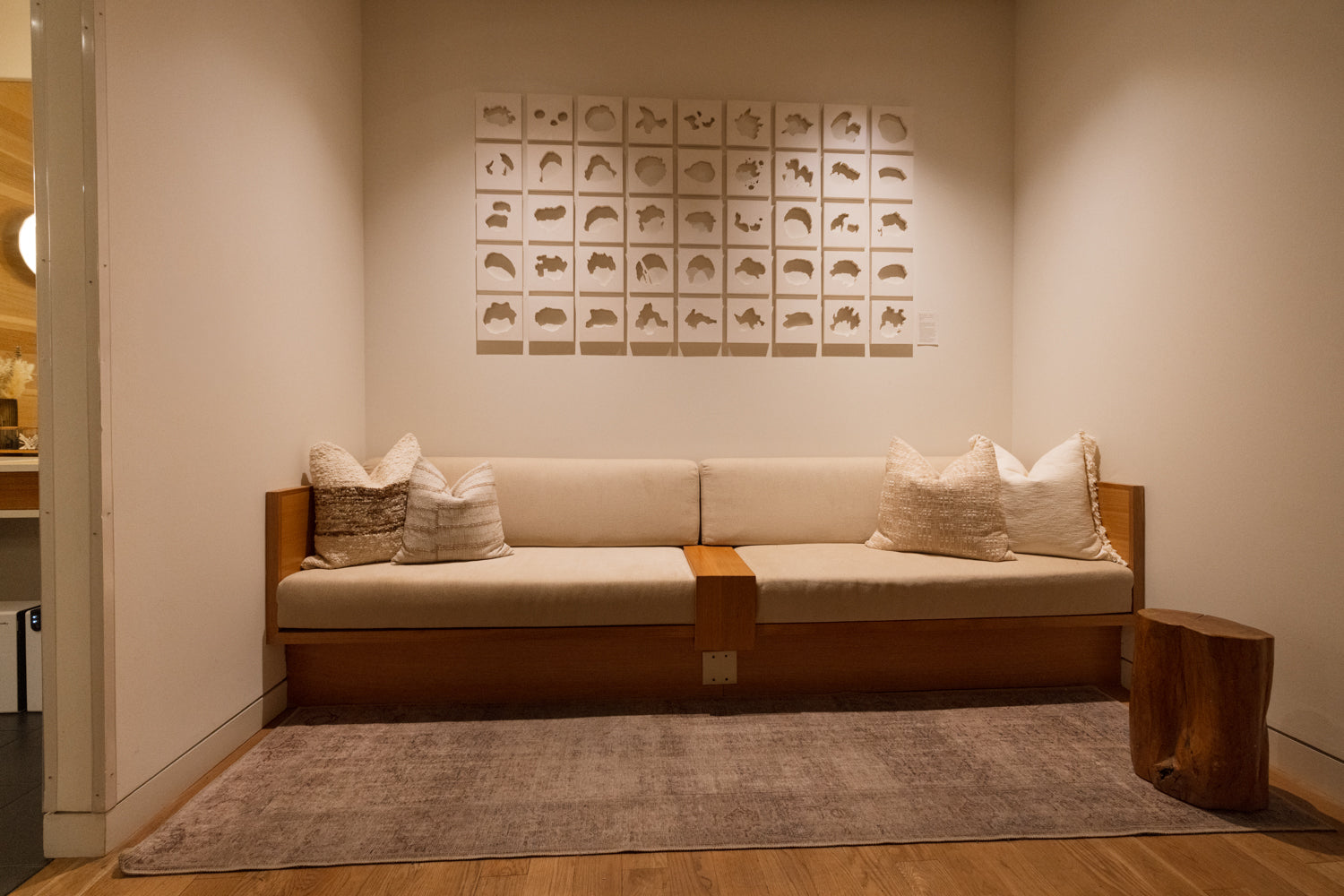 A modern wooden bench with cream cushions and decorative pillows, set against a warm beige wall with a grid of small framed art pieces above. A textured gray rug and a rustic wooden side table are in the foreground.
