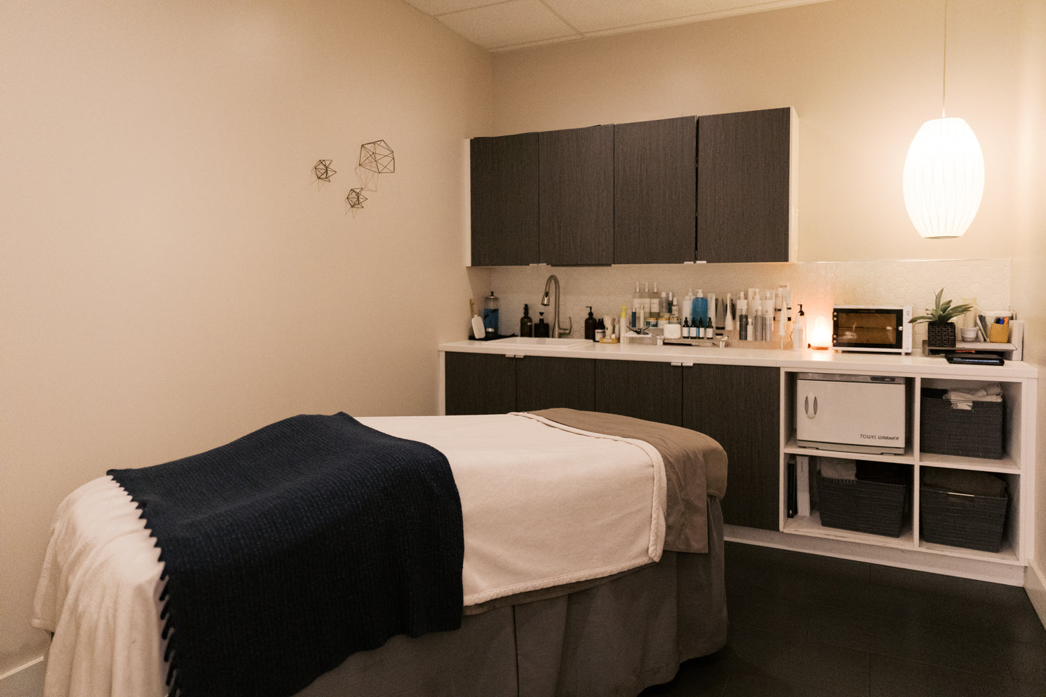 A spa treatment room featuring a massage table with white and navy blue linens, a built-in treatment counter with shelves and cabinets, and a hanging pendant light.
